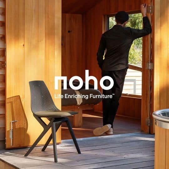 image of wooden home with black chair as the sun hits it. in the background there is a man standing with his back towards you wearing comfortable black attire and grey sneakers. He leans onto the door frame with his feet cross below. Text reads: noho, life enriching furniture