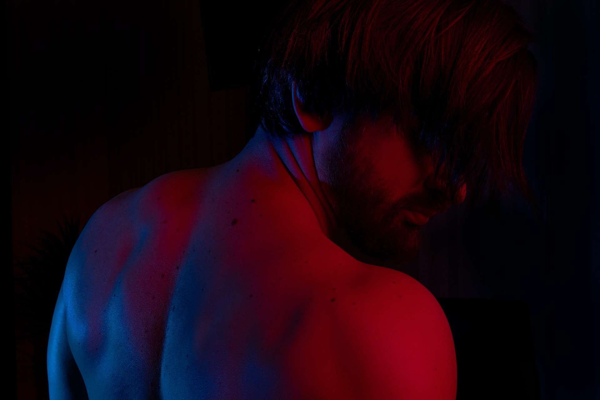 image of a shirtless man with his upper back in frame facing the camera and slightly turned head. He is in a dark space with a red light glowing in his skin.